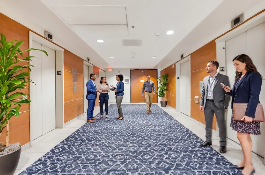 Barrister Suites Office Lease Five people stand and talk in a modern office hallway with elevators on both sides, patterned blue carpet, wooden walls, and potted plants. Two groups converse while one person walks toward the elevators.