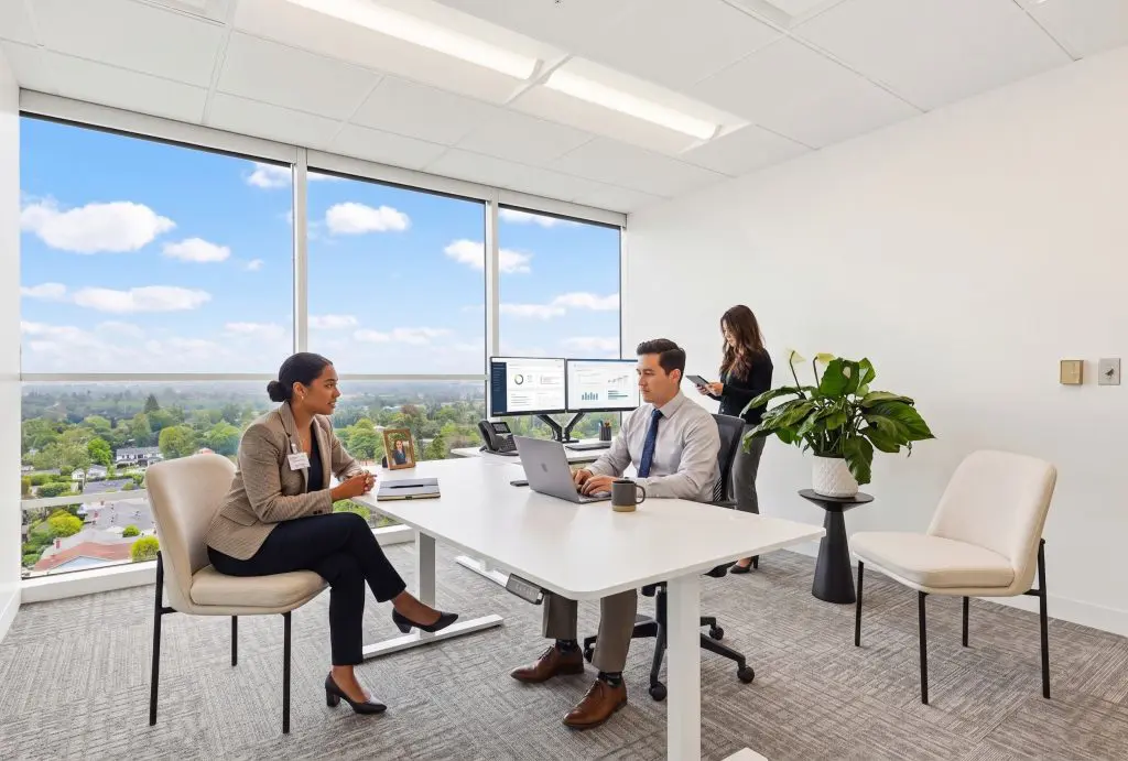 Barrister Suites Office Lease Three professionals in a modern office with large windows; two are seated at a desk talking, with charts on a monitor, while another stands by the window on her phone. A plant sits on the floor by an empty chair.