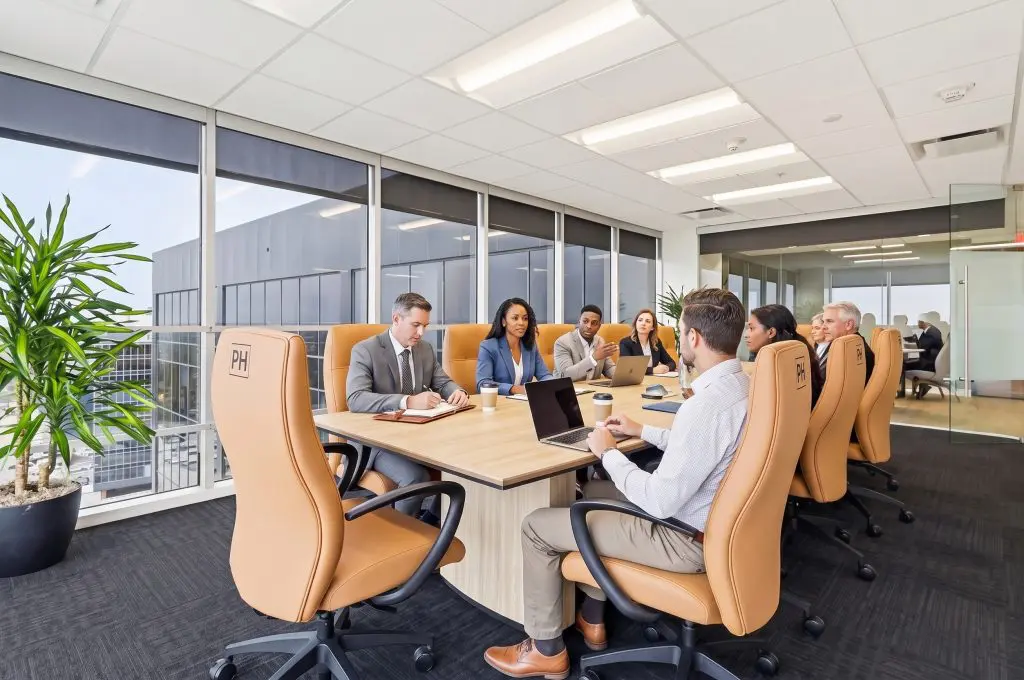 Barrister Suites Office Lease Eight people in business attire are having a meeting in a modern office with large windows, tan chairs, laptops, and notepads on the table.