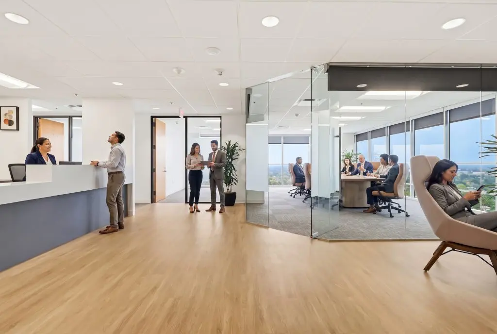 Barrister Suites Office Lease Modern office with people interacting at a reception desk, two colleagues talking in a hallway, a meeting in a glass-walled room, and a person using a smartphone in a lounge chair. Large windows show a city view.