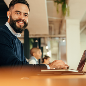 Barrister Suites Office Lease A bearded man in a blue sweater smiles while working on a laptop at a desk in a modern, open office. A takeaway coffee cup sits nearby. Other people work in the background.