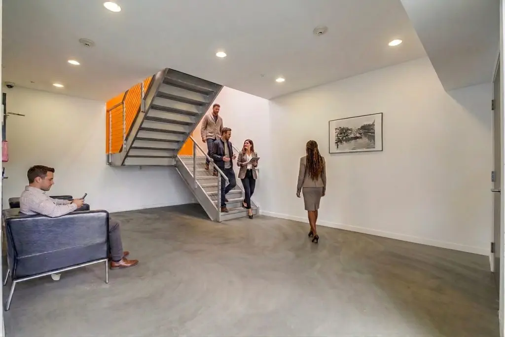 Barrister Suites Office Lease A man sits on a chair in a modern office lobby as three colleagues descend a staircase.