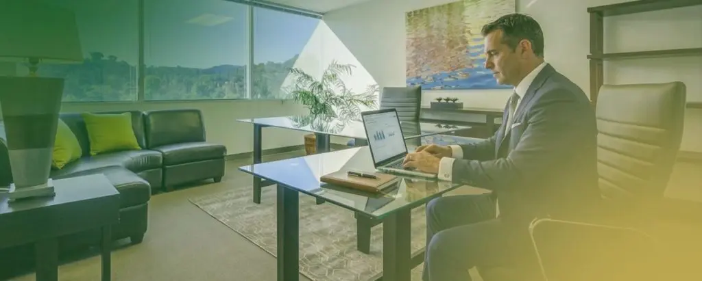 Barrister Suites Office Lease A businessman in a suit sits at a glass desk working on a laptop in a bright office.