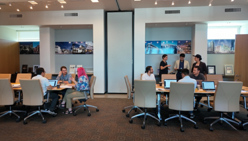 Barrister Suites Office Lease A modern office meeting room with people working in groups at tables, using laptops and having discussions. The room is well-lit, with architectural photos on the walls and beige chairs around the tables.