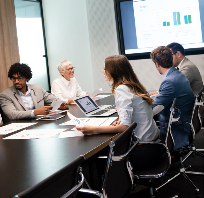 Barrister Suites Office Lease Five people in business attire sit around a conference table with laptops and papers, discussing charts displayed on a large screen in a modern office meeting room.