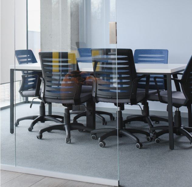 Barrister Suites Office Lease A modern conference room with a glass wall, featuring a white table surrounded by black and blue office chairs on a gray carpeted floor. The room is empty and well-lit by natural light.