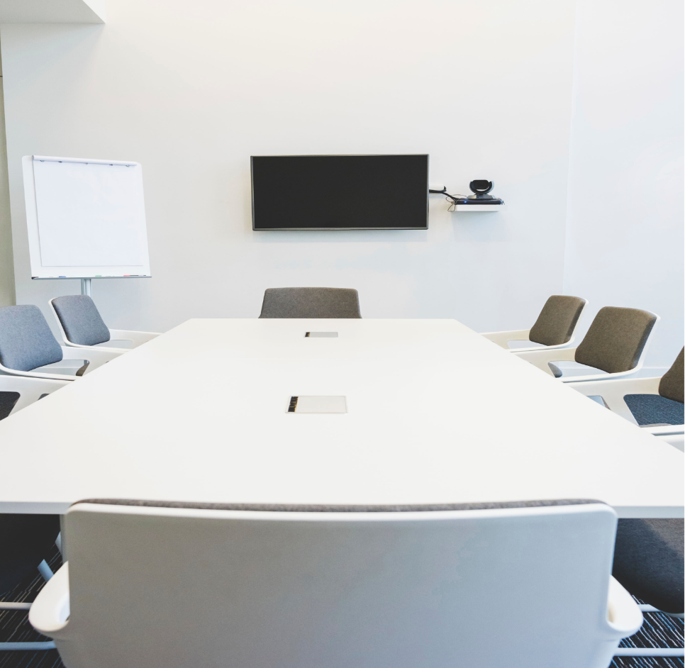 Barrister Suites Office Lease A modern conference room with a long white table surrounded by gray and white chairs, a blank flipchart, and a wall-mounted TV screen with a camera above it. The room has a clean and minimalist design.