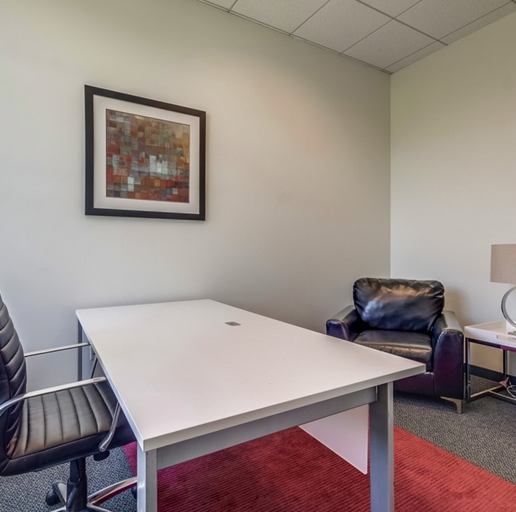 Barrister Suites Office Lease A modern office with a white desk, a black leather office chair, a black armchair, a table lamp on a side table, and abstract artwork on a cream wall above a red rug.
