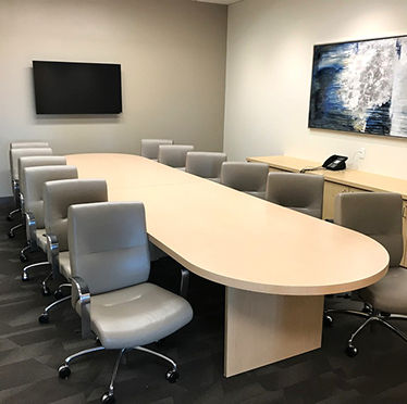 Barrister Suites Office Lease A modern conference room with a large oval wooden table surrounded by twelve gray office chairs, a wall-mounted TV, a telephone on a side cabinet, and an abstract painting on the wall.