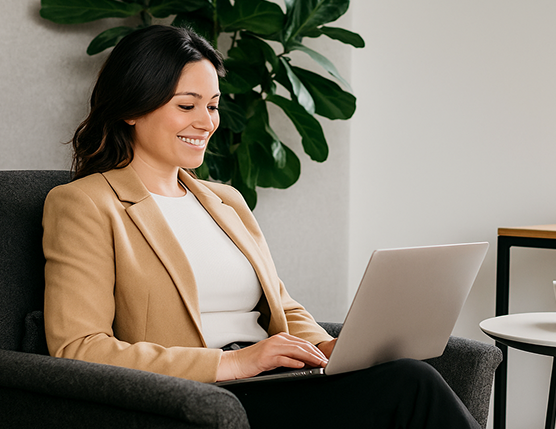 Barrister Suites Office Lease A woman in a tan blazer and white top sits in a chair, smiling while typing on a laptop. A green plant and modern furniture are visible in the background.
