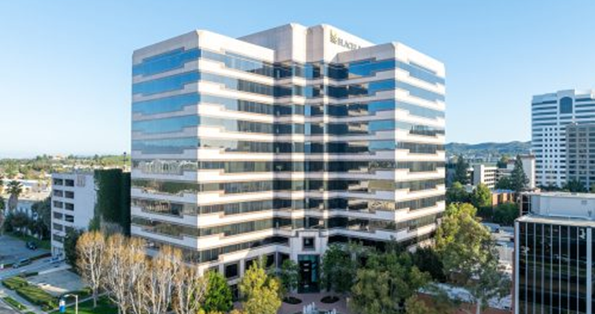 Barrister Suites Office Lease A large, modern office building with reflective glass windows and multiple stories stands surrounded by trees and smaller buildings on a clear, sunny day.