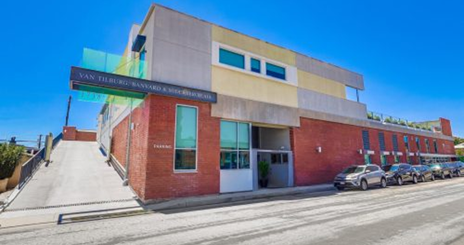 Barrister Suites Office Lease A modern two-story building with red brick and beige walls, large windows, and a sign reading “Van Tilburg, Banvard & Soderbergh.” Several parked cars line the street in front under a clear blue sky.