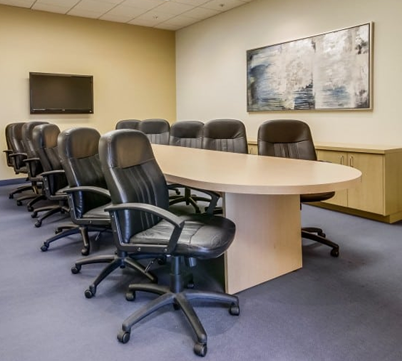 Barrister Suites Office Lease A modern conference room with a long oval wooden table, surrounded by eleven black leather office chairs. A flat-screen TV is mounted on the wall and an abstract painting hangs above a light wood cabinet.