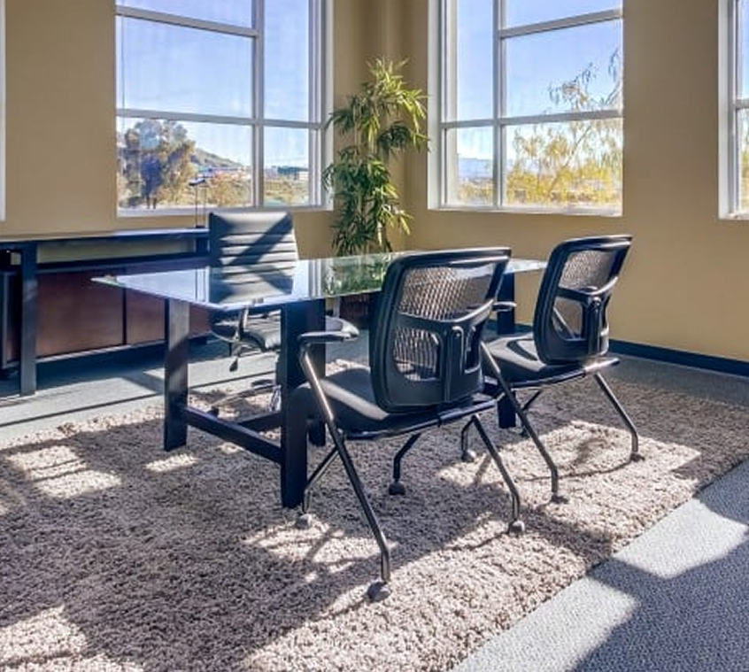 Barrister Suites Office Lease A modern office with a glass table, three black mesh chairs, large windows letting in sunlight, a shaggy carpet, and a tall plant in the corner.