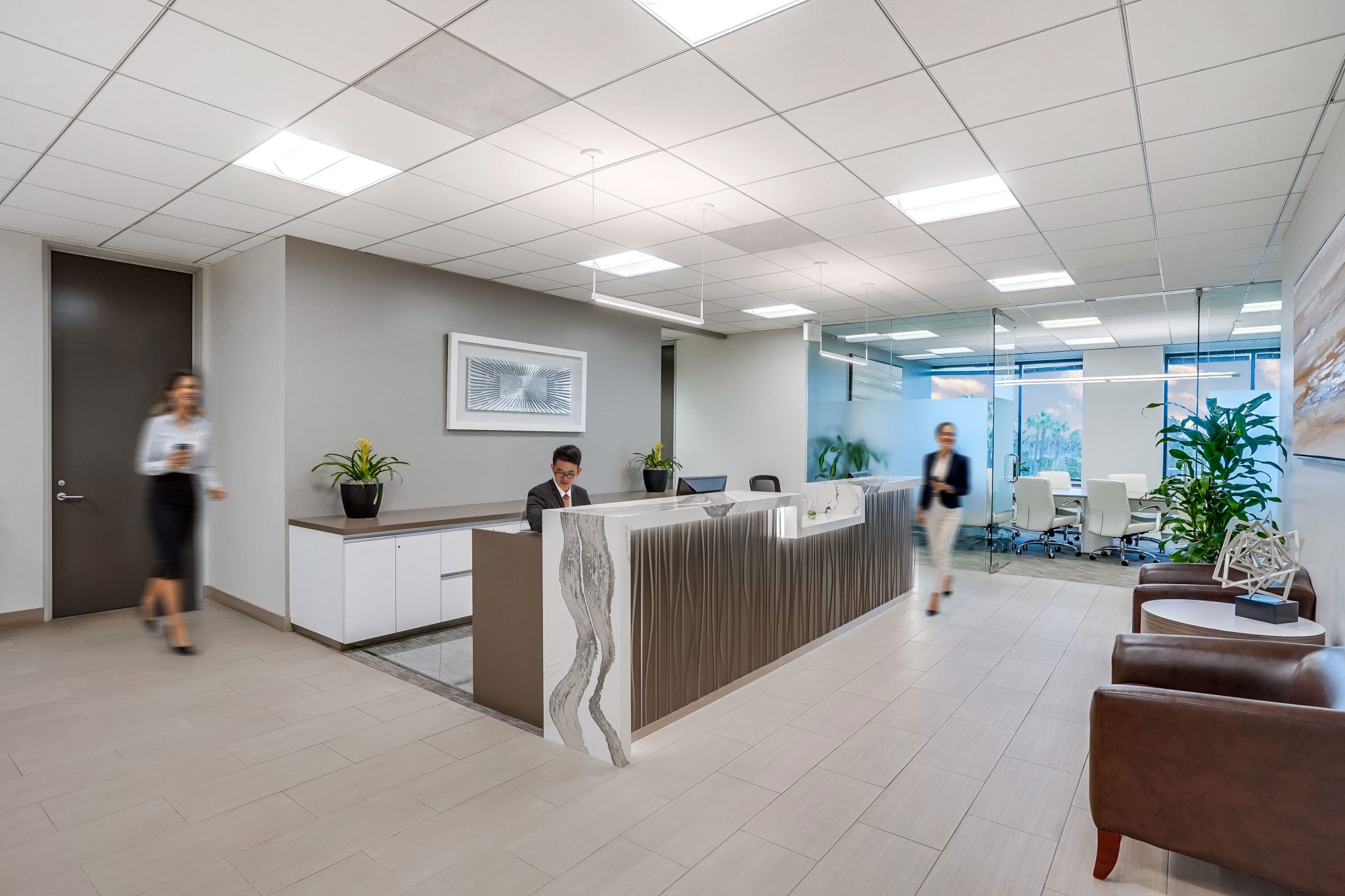 Barrister Suites Office Lease Modern office lobby with a reception desk, receptionist working, two people walking, potted plants, brown chairs, and a conference room with glass walls in the background. The space is well-lit and decorated.