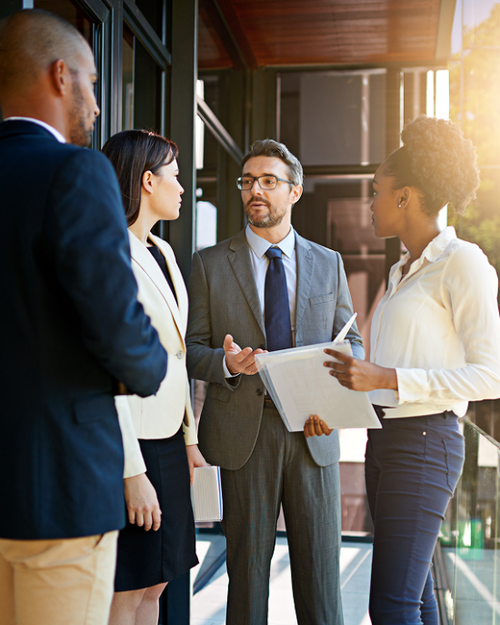 Barrister Suites Office Lease Four business professionals stand outside a glass building, engaged in conversation. One man in a suit speaks while holding papers; the others listen attentively. Sunlight shines from the right side of the image.