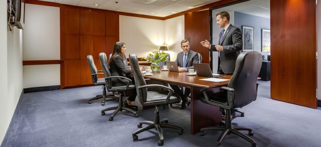 Barrister Suites Office Lease Three business professionals in formal attire meet in a conference room; two are seated at a long table with a laptop, while one stands and speaks. The room has wood paneling and office chairs.