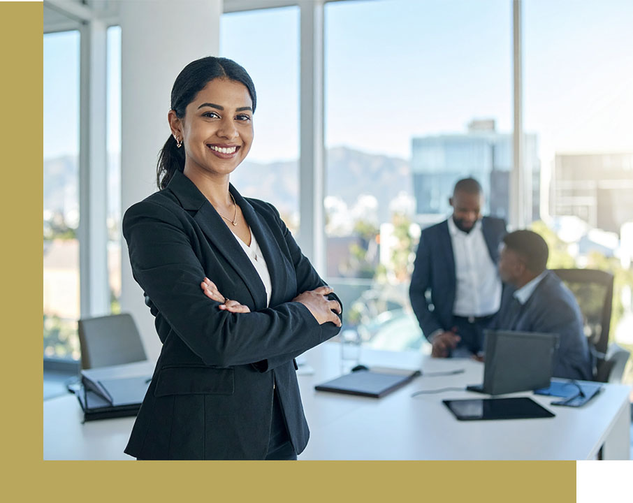 Barrister Suites Office Lease A confident businesswoman in a suit stands with folded arms in a bright office, smiling at the camera. Two colleagues are having a discussion in the background near a window with city views.