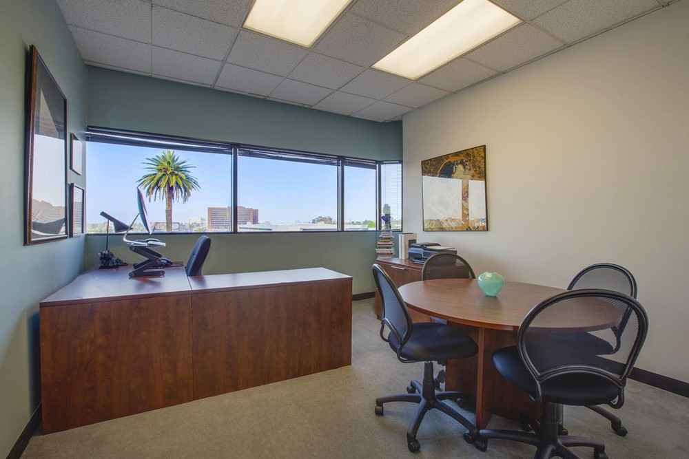 Barrister Suites Office Lease Modern office with a wooden desk, computer, three chairs around a round table, framed art on the walls, and large windows overlooking a cityscape with a palm tree.