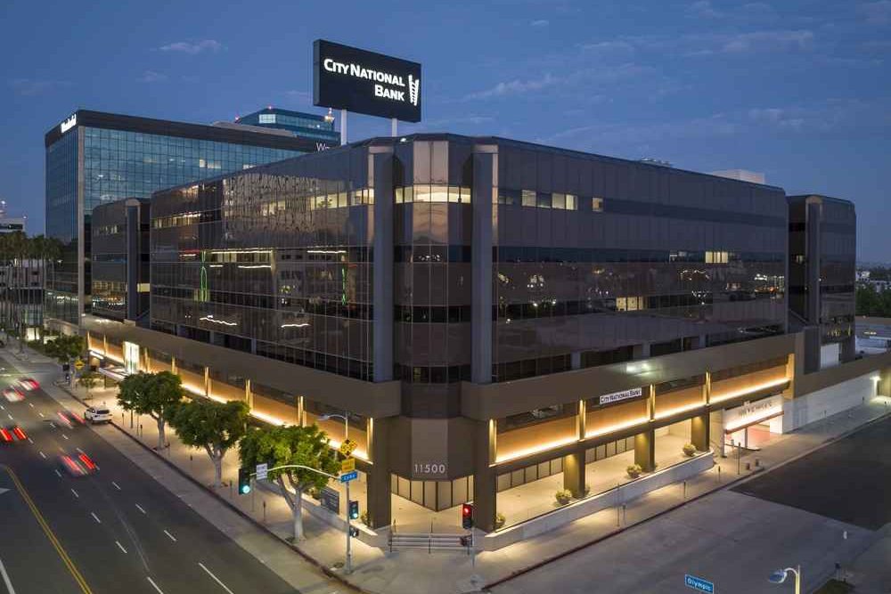Barrister Suites Office Lease A modern, multi-story office building at a city intersection, illuminated at dusk, with City National Bank signage on the rooftop and glass windows reflecting the evening sky.