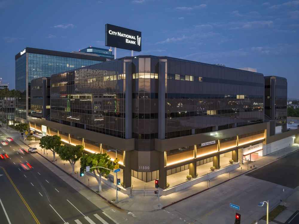 Barrister Suites Office Lease A modern, multi-story office building at a city intersection, illuminated at dusk, with City National Bank signage on the rooftop and glass windows reflecting the evening sky.