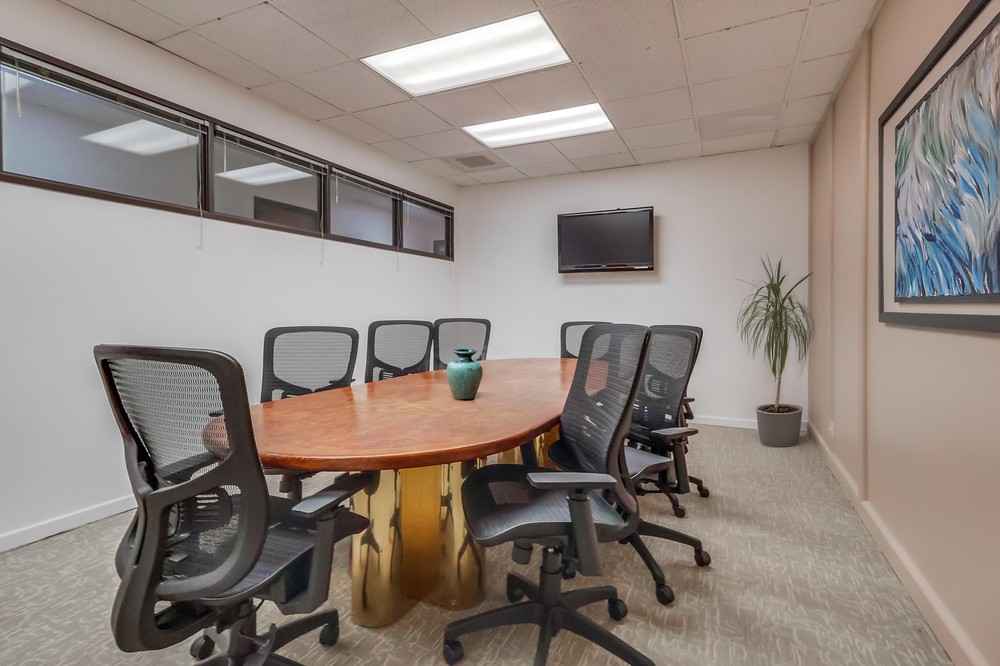 Barrister Suites Office Lease A modern conference room with an oval wooden table, eight black office chairs, a wall-mounted TV, a potted plant, and a framed abstract painting on the wall. Fluorescent ceiling lights illuminate the room.