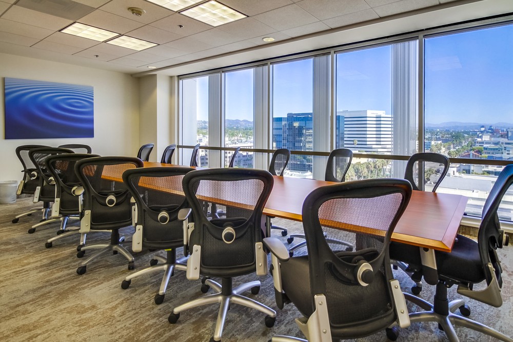 Barrister Suites Office Lease A modern conference room with a long wooden table surrounded by black mesh office chairs, large windows letting in natural light, and a cityscape view outside. A blue abstract painting hangs on the wall.