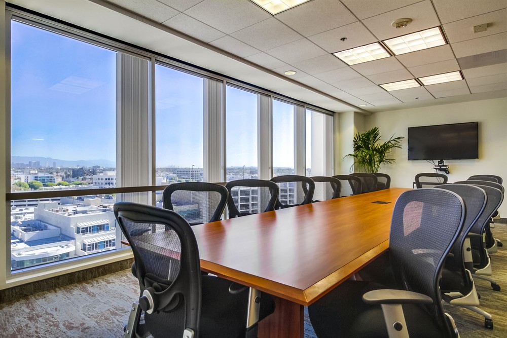 Barrister Suites Office Lease A modern conference room with a long wooden table, black mesh chairs, large windows with a city view, a flat-screen TV on the wall, and a potted plant in the corner.