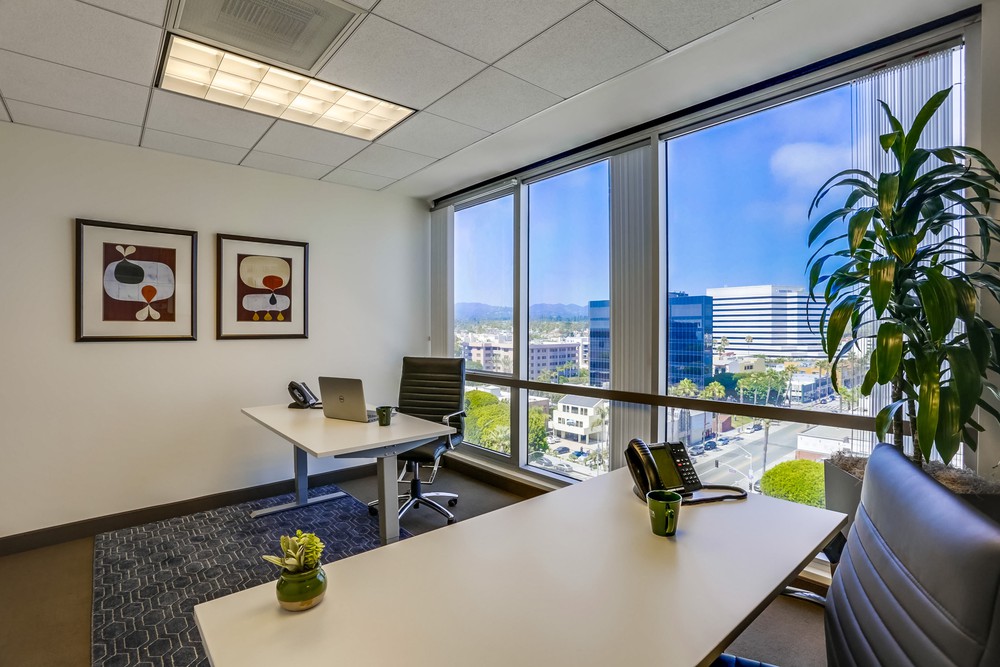 Barrister Suites Office Lease A modern office with two desks, chairs, a laptop, a phone, and plants. Large windows offer a cityscape view with buildings and blue sky. Two abstract artworks hang on the wall.