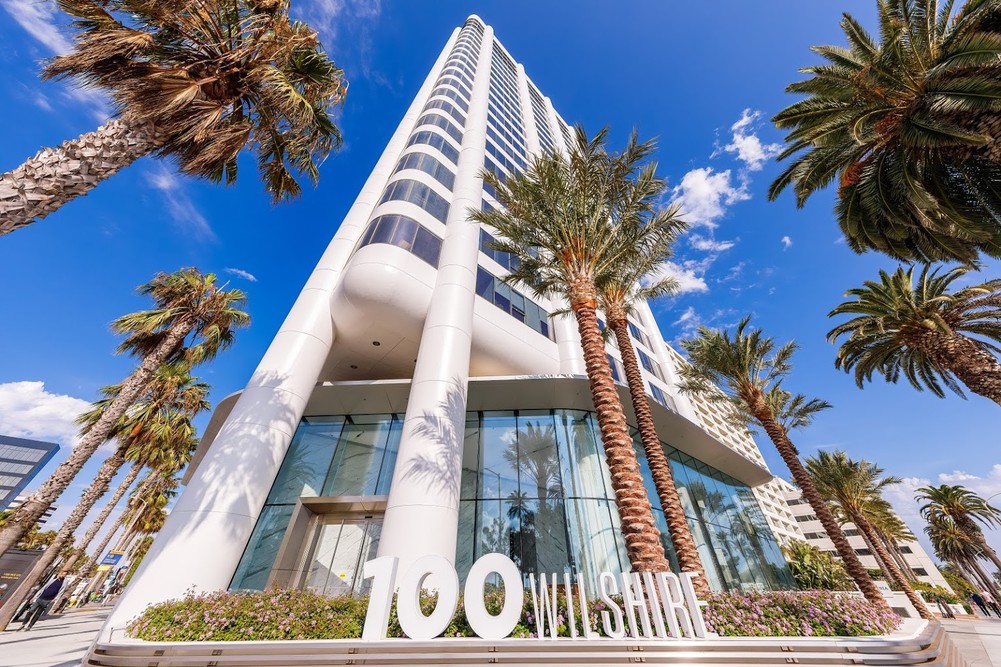 Barrister Suites Office Lease A tall white office building with curved corners and large glass windows is surrounded by palm trees. The address 100 Wilshire is displayed in large letters in front. The sky is blue with scattered clouds.