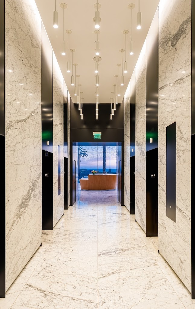 Barrister Suites Office Lease A modern hallway with marble floors and walls, multiple ceiling lights, and black elevator doors. At the end, there is a seating area with a window view of palm trees and a sunset.