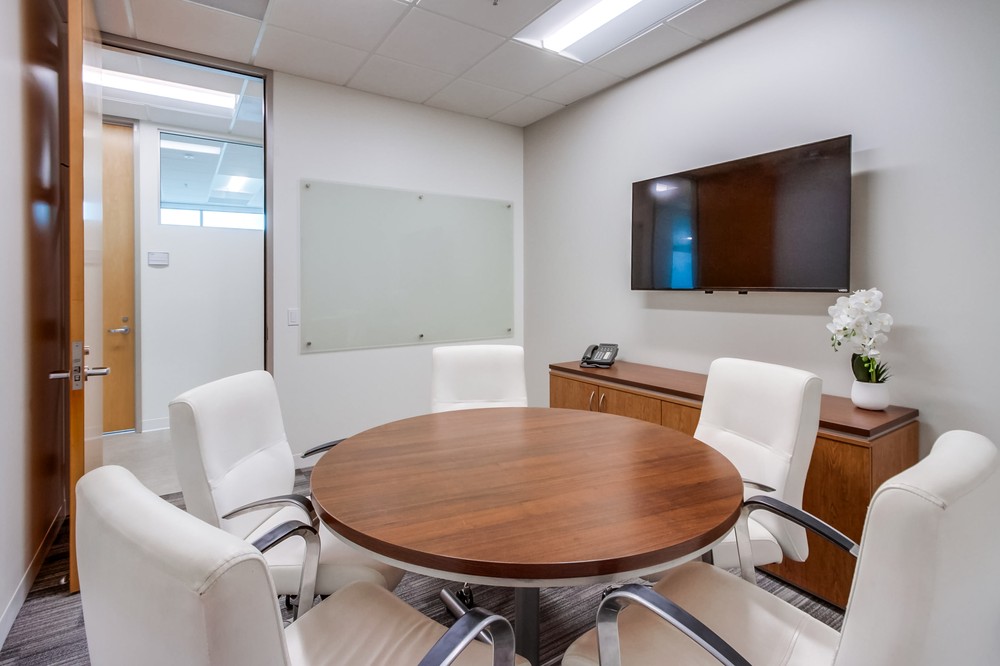Barrister Suites Office Lease A small modern conference room with a round wooden table, five white chairs, a wall-mounted TV, a whiteboard, and a cabinet with a phone and a potted plant. The room has neutral walls and bright overhead lighting.