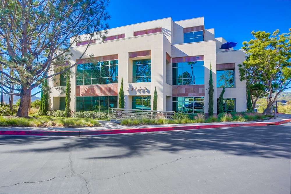 Barrister Suites Office Lease A modern three-story office building with large reflective windows, surrounded by trees and landscaping, under a clear blue sky. The building sits beside a wide, paved road with a red curb.