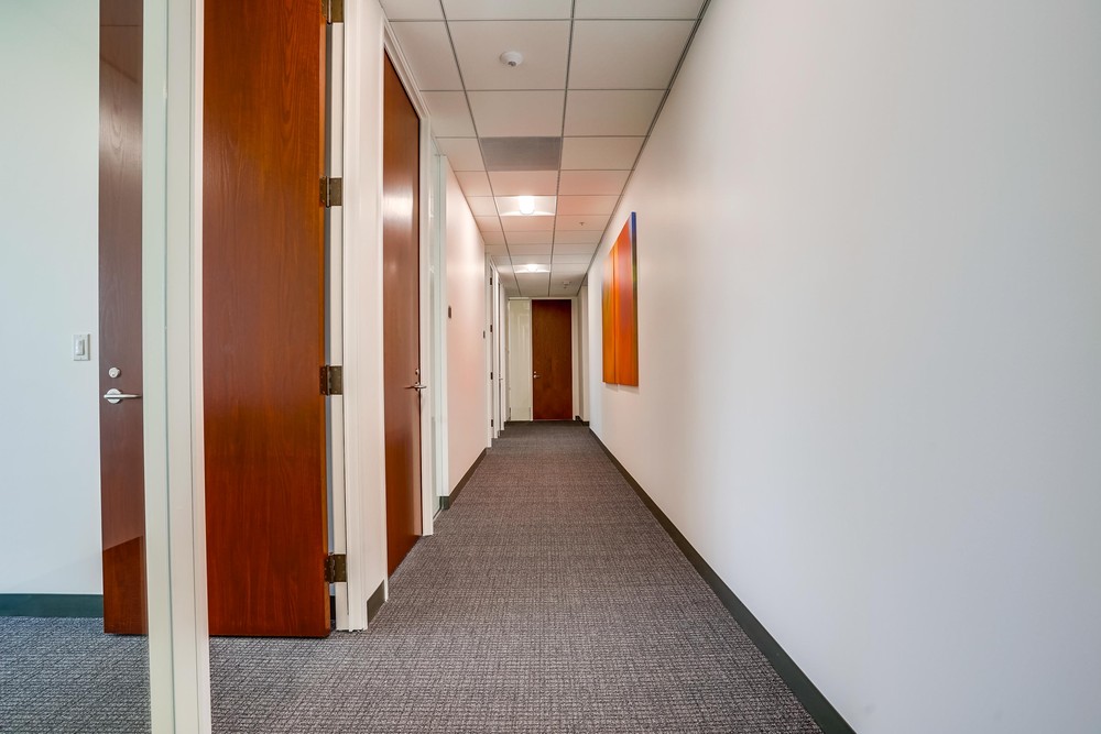 Barrister Suites Office Lease A clean, carpeted office hallway with several closed wooden doors on the left, white walls, a ceiling with recessed lighting, and a colorful abstract painting on the right wall.
