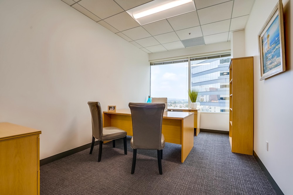 Barrister Suites Office Lease A modern office with two beige chairs facing a wooden desk, set near a large window with a city view. The room has white walls, carpeted floor, and a framed painting on the right wall.