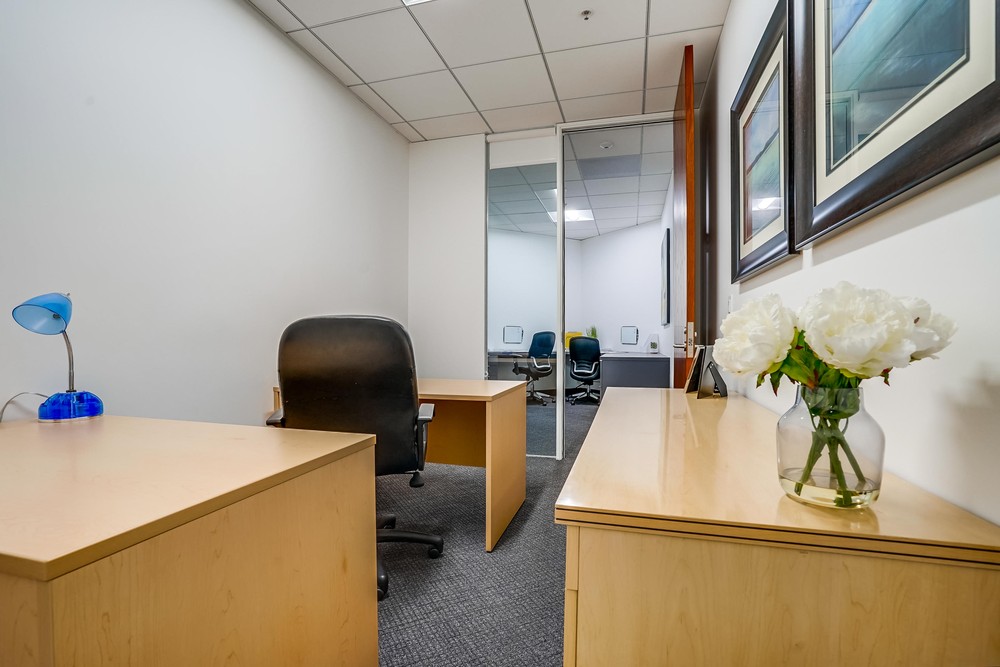 Barrister Suites Office Lease A small modern office with light wood desks, black chairs, framed art on the walls, a blue desk lamp, and a vase of white flowers. A glass divider separates the space from another office area.