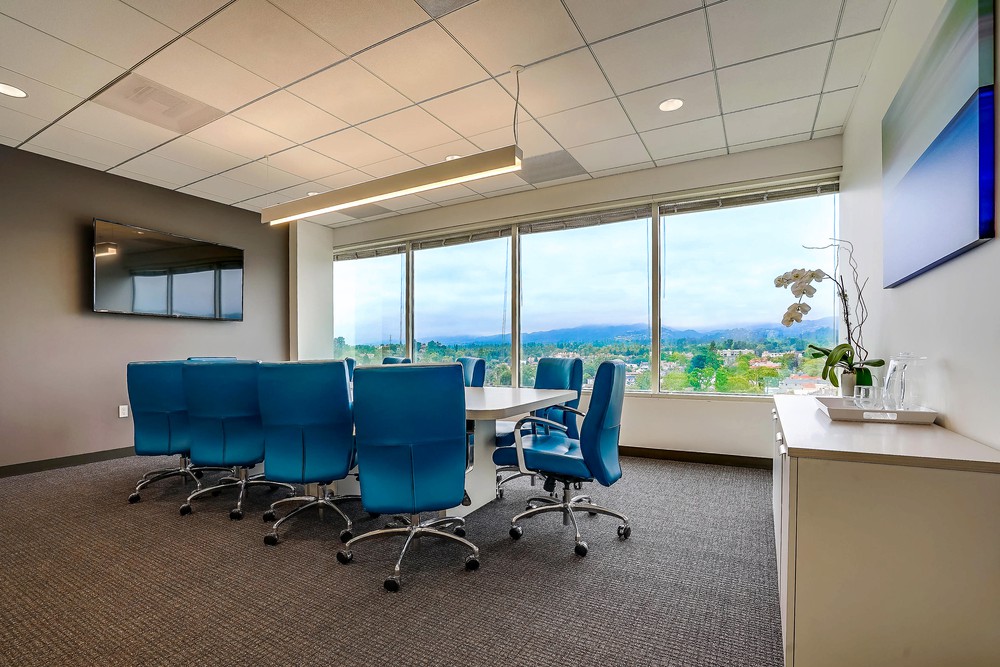 Barrister Suites Office Lease Modern conference room with a large table surrounded by blue chairs, a wall-mounted TV, and large windows offering a scenic view of trees and mountains. A cabinet with glasses and a plant is on the right.