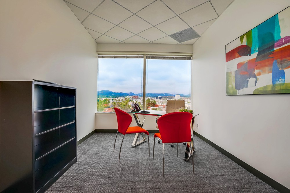 Barrister Suites Office Lease A small office with two red chairs at a glass table, a black bookshelf, large window with a city and mountain view, and a colorful abstract painting on the wall.