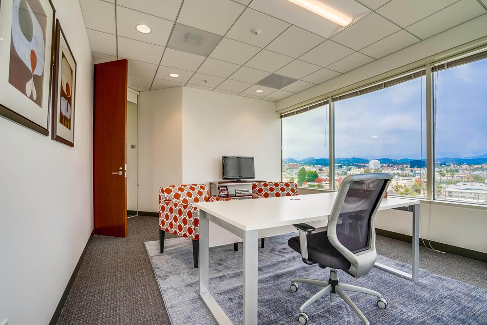 Barrister Suites Office Lease Modern office with a white desk, mesh chair, small TV, and a sofa with red and white pattern. Large windows offer a city view; two abstract art pieces hang on the wall. Door is open, letting in natural light.