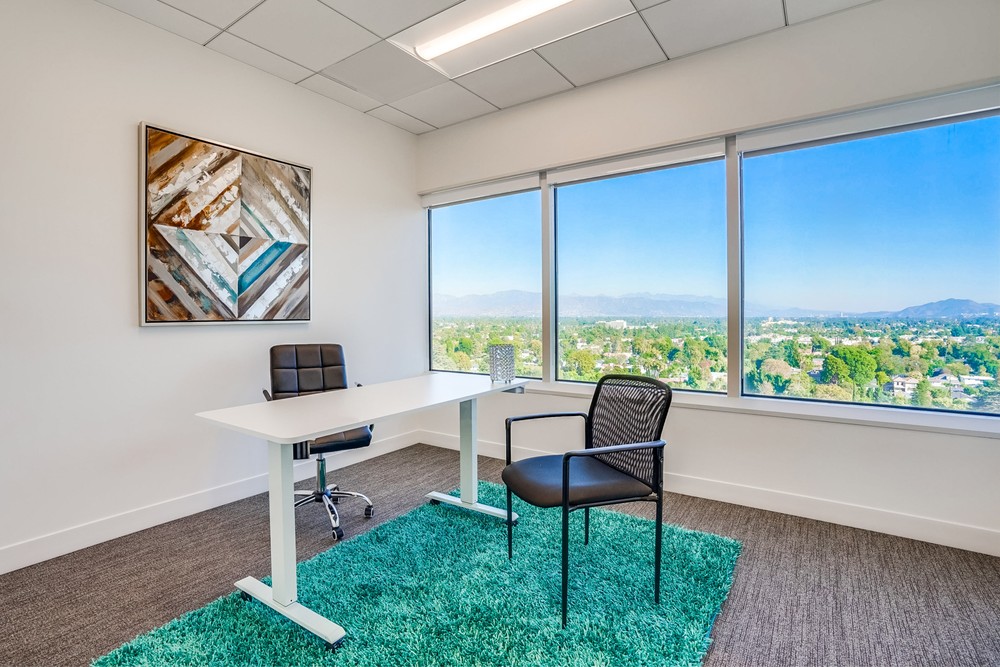 Barrister Suites Office Lease A modern office with a white desk, two chairs, a green rug, abstract wall art, and large windows showcasing a scenic city and mountain view under a clear blue sky.