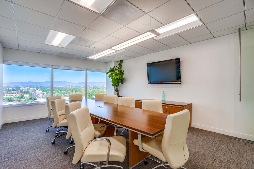 Barrister Suites Office Lease A modern conference room with a wooden table, eight white chairs, a wall-mounted TV, a potted plant, and large windows offering a scenic city and mountain view.