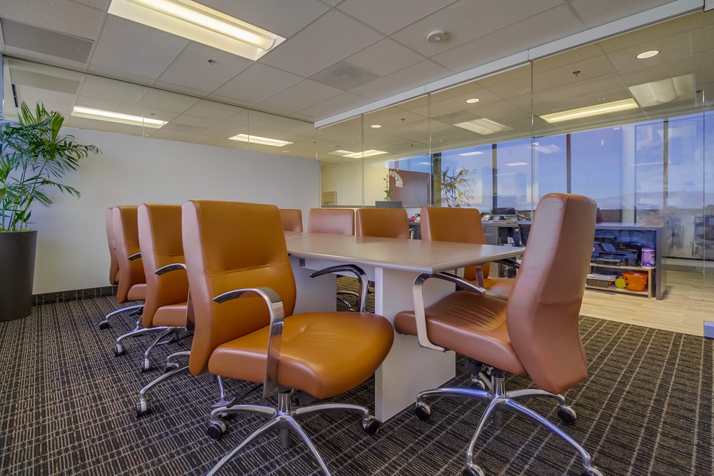 Barrister Suites Office Lease A modern conference room with a long table surrounded by eight brown leather chairs on wheels, set on a carpeted floor, with glass walls and a potted plant in the corner.