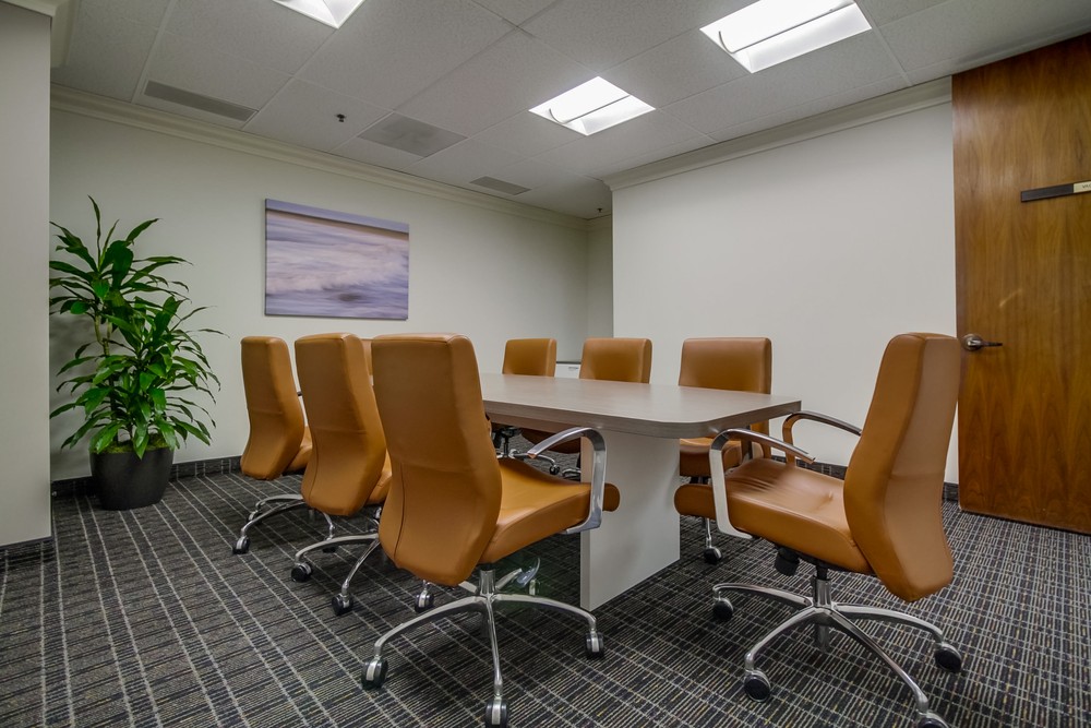 Barrister Suites Office Lease A small conference room with eight tan leather chairs around a rectangular table, a potted plant in the corner, a door, and a framed picture of waves on the wall. The room is brightly lit with overhead lights.