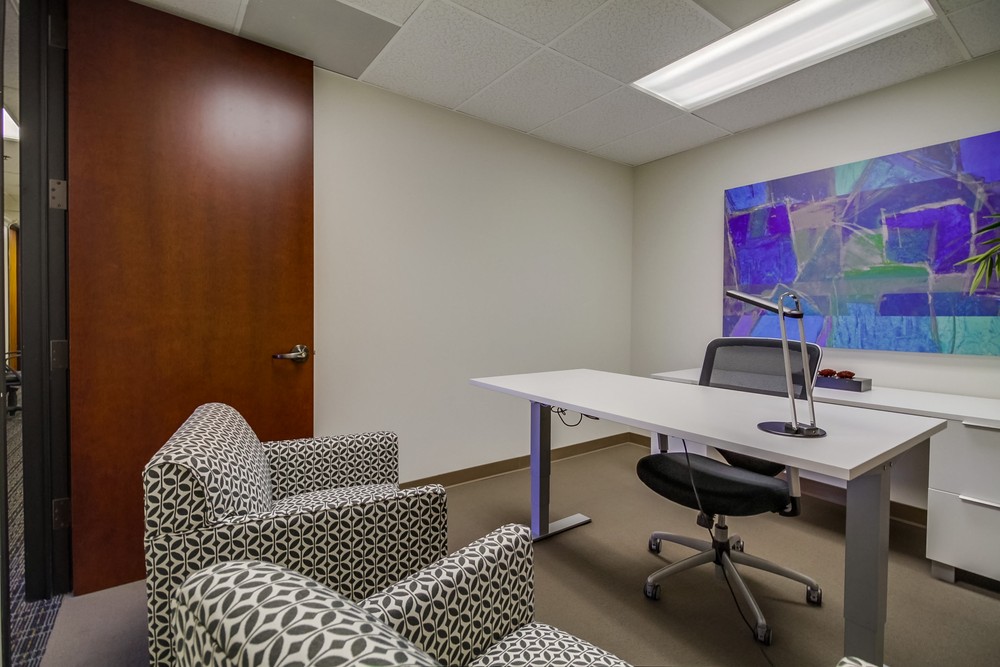 Barrister Suites Office Lease A modern office with a white desk, black office chair, two patterned armchairs, a lamp, and colorful abstract art on the wall. The room has neutral walls and ceiling with fluorescent lighting.