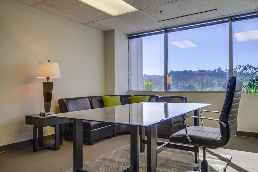 Barrister Suites Office Lease Modern office with a glass desk, black swivel chair, black leather sofa with green pillows, a side table with a lamp, large window with a scenic outdoor view, and potted plant in the corner.