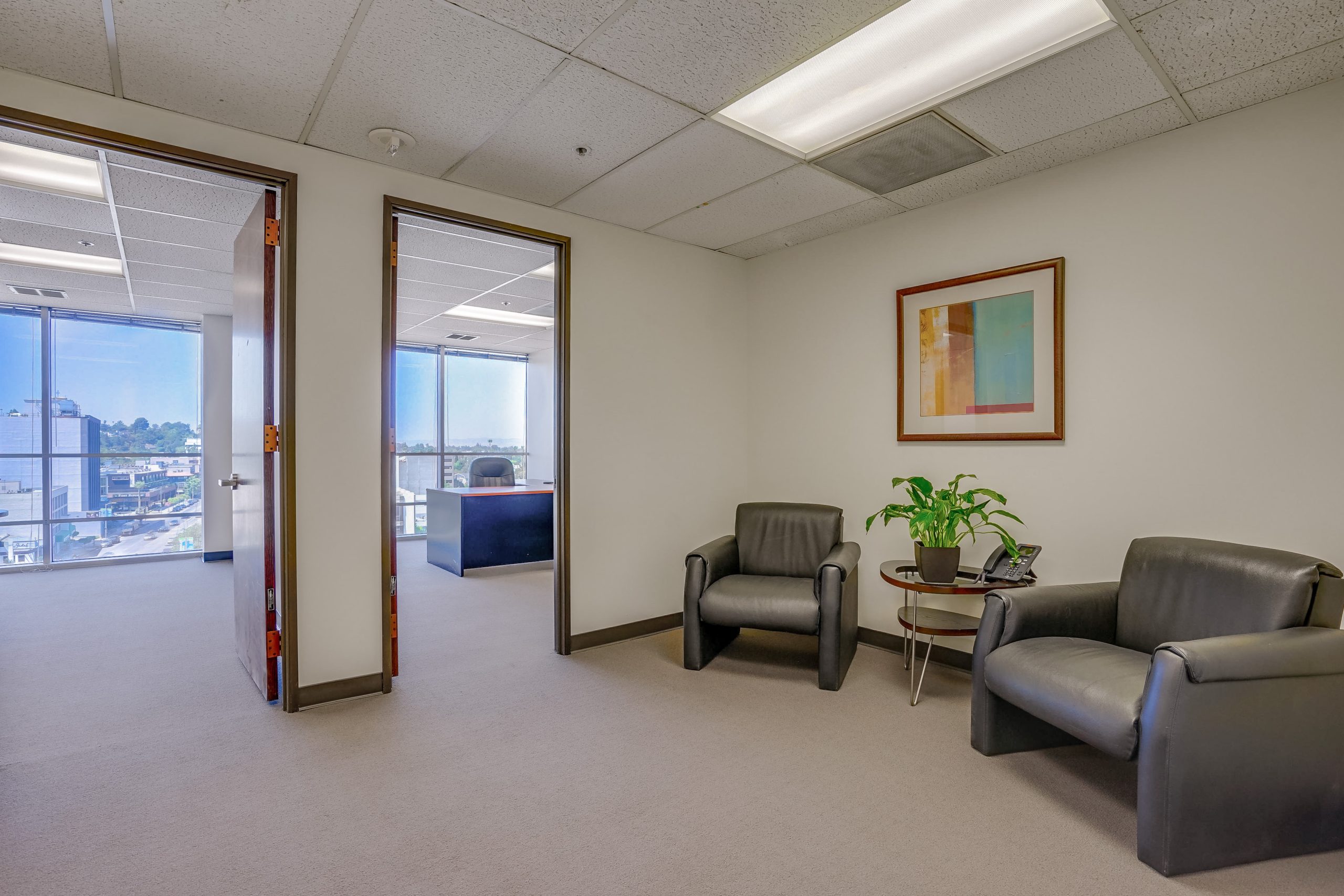 Barrister Suites Office Lease A small office waiting area with two black chairs, a round glass table with a green plant, and a colorful framed artwork on the wall. Open doors reveal an adjoining office with large windows and a city view.