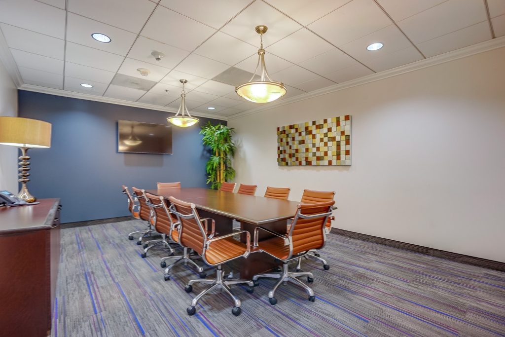 Barrister Suites Office Lease A modern conference room with a long table surrounded by orange office chairs, a wall-mounted TV, abstract wall art, potted plant, and overhead lighting. The room has a carpeted floor and white ceiling tiles.