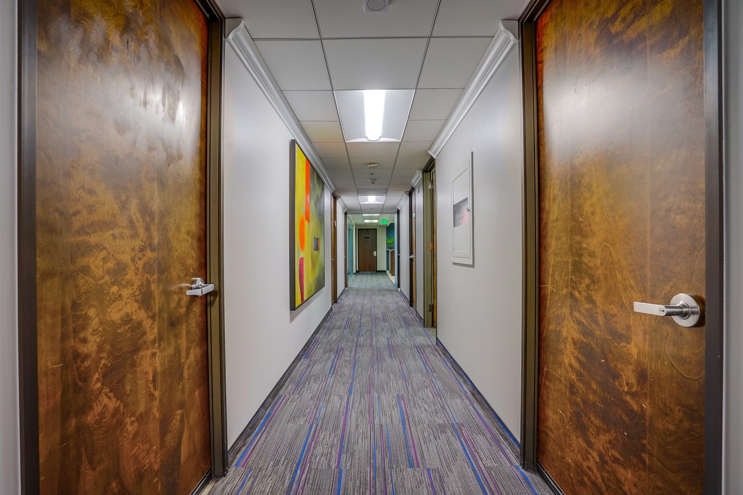 Barrister Suites Office Lease A brightly lit hallway with wooden doors on both sides, colorful abstract artwork on the left wall, and a blue-gray carpet leading to another door at the far end.