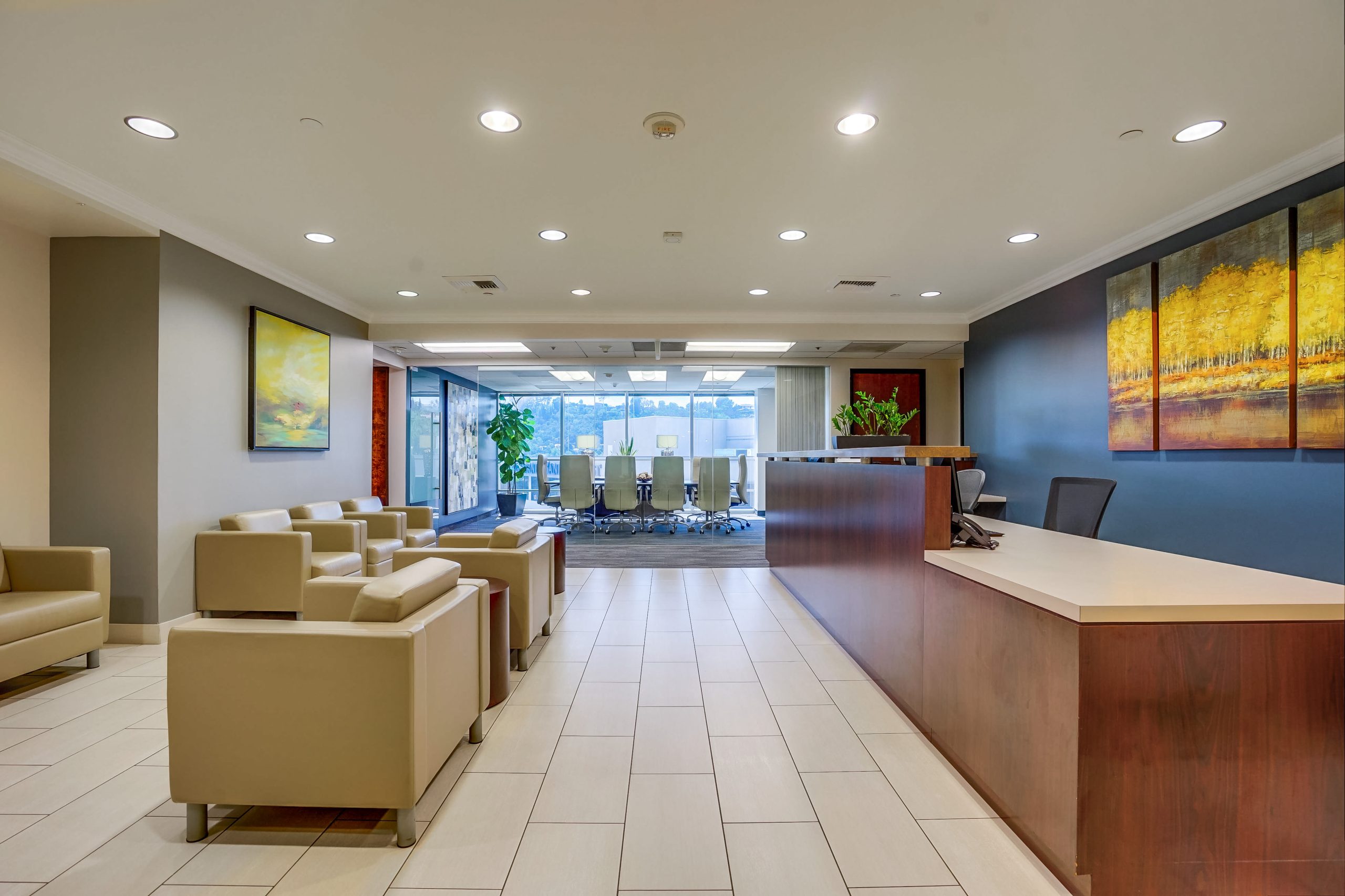 Barrister Suites Office Lease Modern office lobby with beige armchairs, a wooden reception desk, tile flooring, colorful wall art, and large windows in the background revealing a conference room with a view outside.