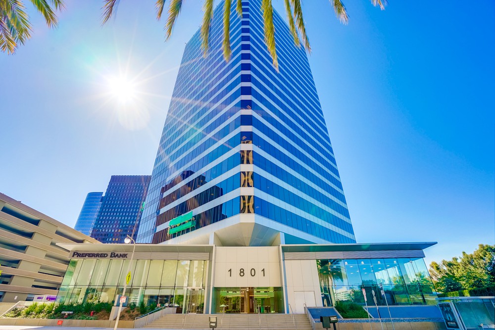 Barrister Suites Office Lease A modern glass high-rise building with 1801 and Preferred Bank at the entrance, viewed from below on a sunny day with palm tree leaves and a bright sun in the blue sky above.
